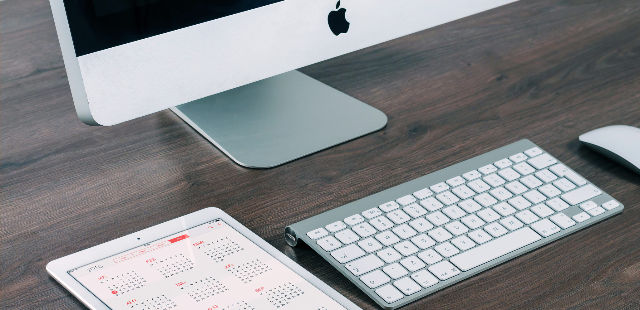 Aufgeräumter Arbeitsplatz mit iMac, Tastatur, Maus und iPad mit Kalender auf einem Holztisch – Webdesign von SHIWA.DE